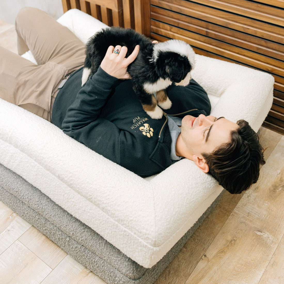 Person lying on 2 white and grey stacked luxury orthopedic dog beds holding a small black and white puppy.