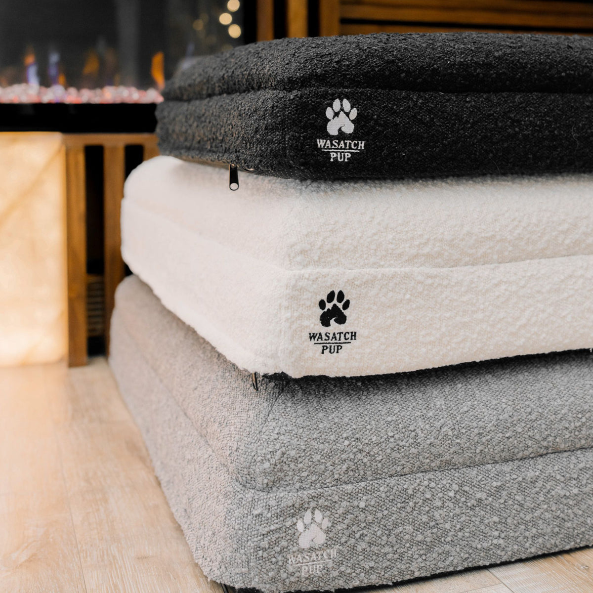 Three Wasatch Pup Luxury Orthopedic Dog Beds stacked in a home setting, showing Charcoal, Grey, and Bone bouclé fabric options. Each rectangular bed has a plush removable cover, cushioned sleeping surface, and raised foam bolsters. The orthopedic construction includes memory foam, high-density support foam, waterproof protective liners, and a non-slip bottom.